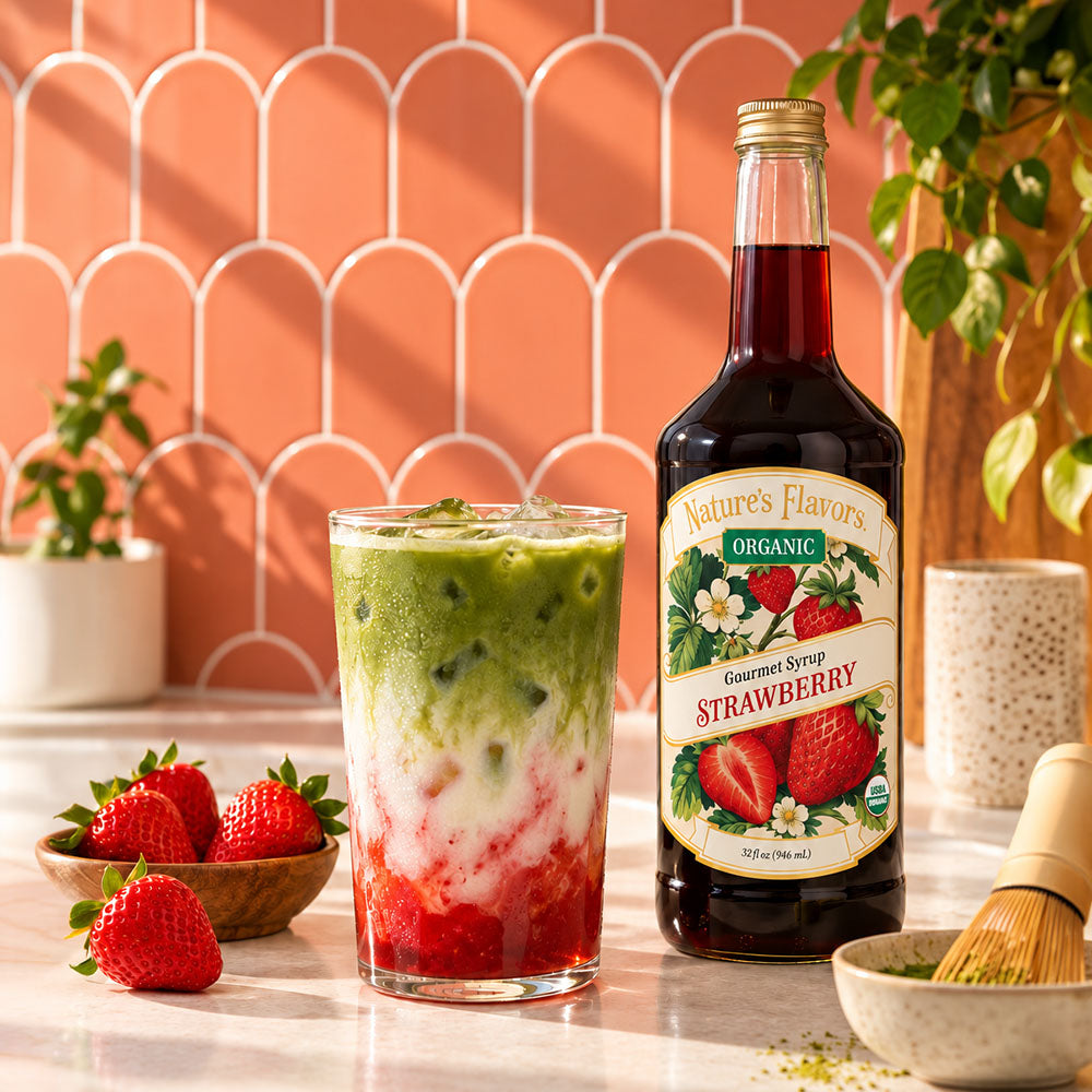 A bottle of nature's Flavors Organic Strawberry Syrup Stands next to a strawberry matcha latte in a kitchen with peach tiles and greenery.
